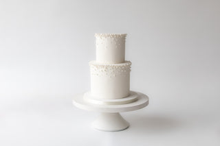 Two-tiered white cake with decorative elements on a white stand against a white background
