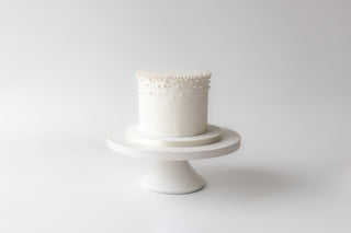 White cake with decorative top on a white stand against a light gray background