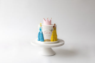 Cake with princess figures and a pink crown on a white stand against a white background