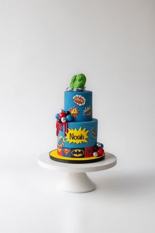 Colorful superhero-themed cake on a white stand against a white background