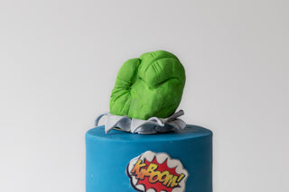 Green plush toy on a blue cake with 'K-BOOM!' decoration against a white background