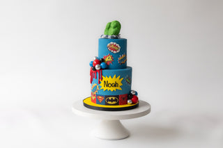 Two-tiered birthday cake with superhero-themed decorations on a white stand against a white background